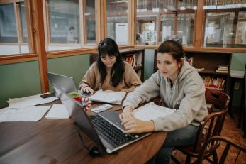 Two people sit at a table and work on laptops.