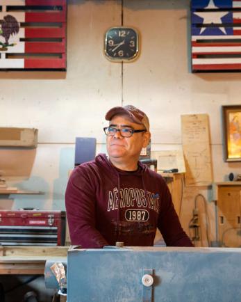 owner of woodworking shop stands below a Mexican and American flag