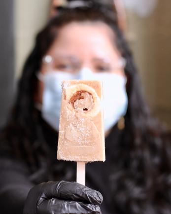 woman with black hair and mask holds a cream colored popsicle that she made
